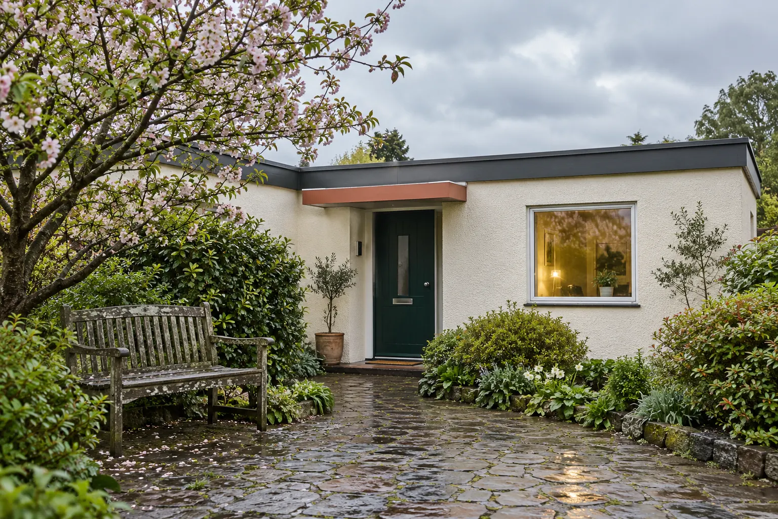 Cremefarbenes Flachdachhaus mit dunkelgrüner Tür, Terrakottavordach und beleuchtetem Fenster hinter nassem Gartenpflaster und Holzbank. KI-generiertes Motiv.