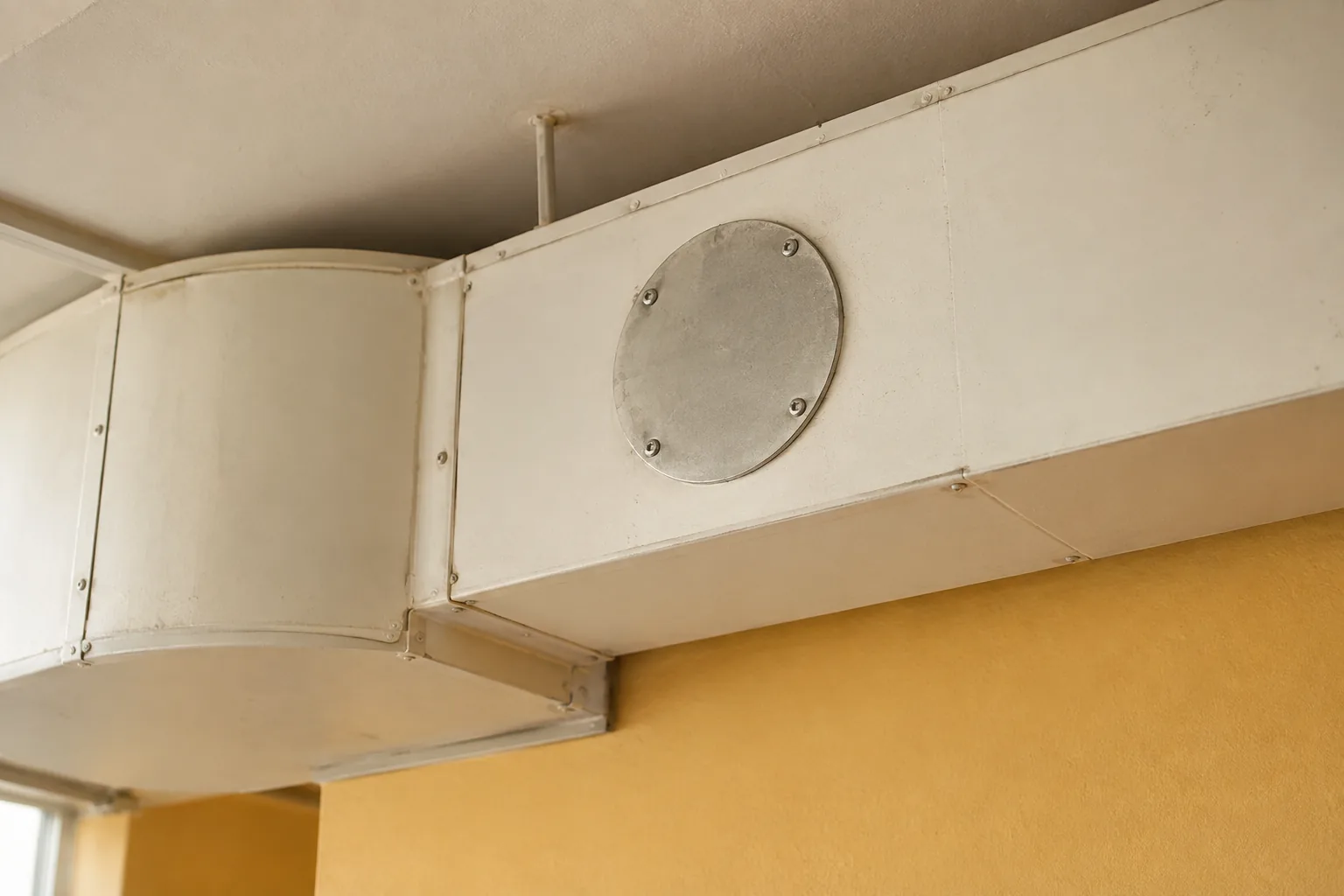 Vollständig geschlossener runder Metalldeckel mit sichtbaren Schrauben an einem weiß gestrichenen rechteckigen Lüftungskanal über einer ockerfarbenen Innenwand. KI-generiertes Detailmotiv.