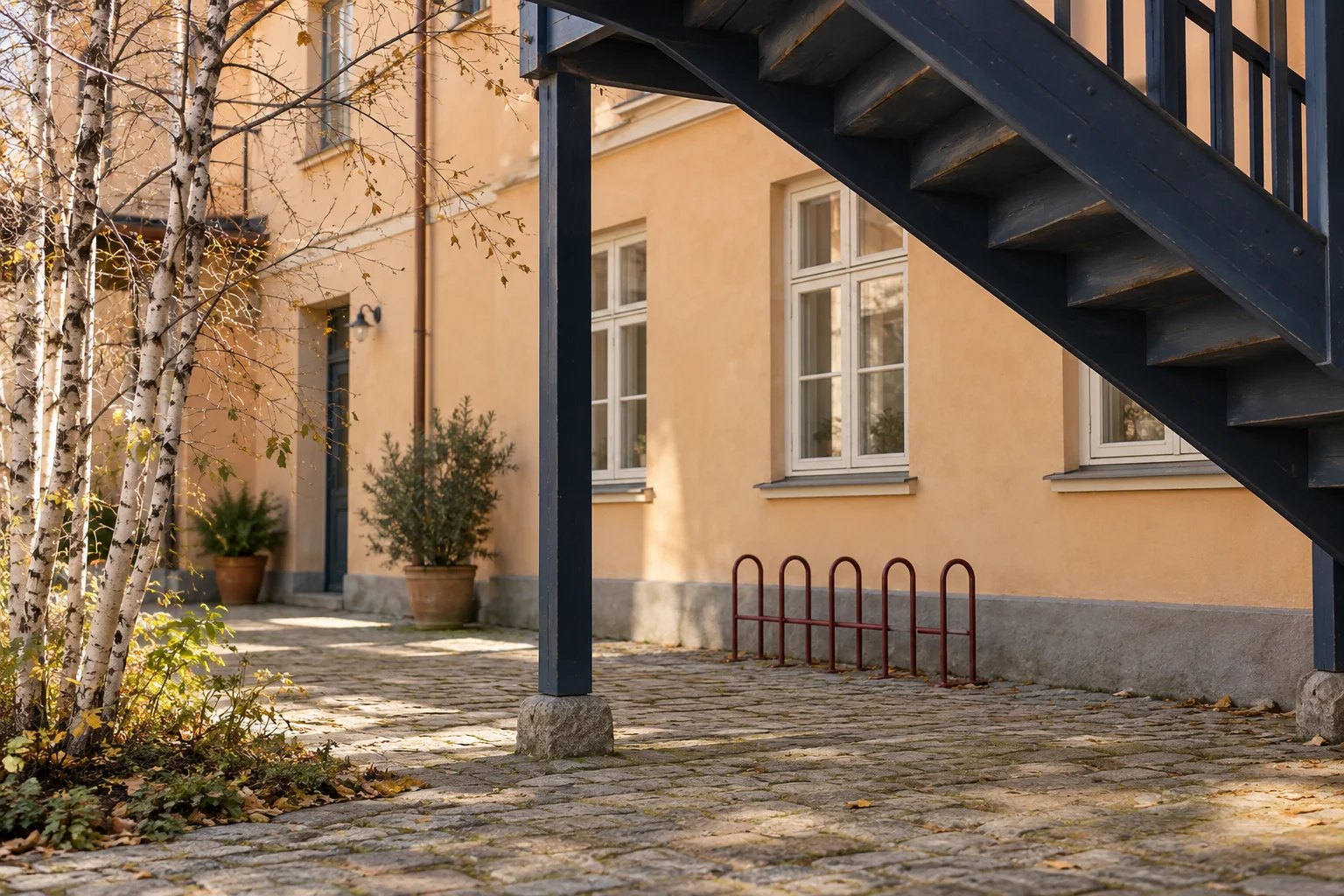Eine dunkelblaue hölzerne Außentreppe überspannt den Hof vor einer apricotfarbenen Fassade mit geschlossenen hellen Fenstern, leerem roten Fahrradständer und Birken.