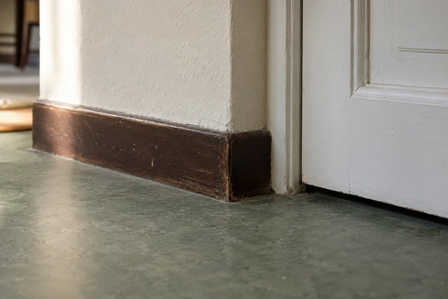 Nahansicht eines cremefarbenen Wandabschlusses mit dunkler Holzsockelleiste, grünlichem Bodenbelag und geschlossenem weißem Türblatt. KI-generiertes Detailmotiv.